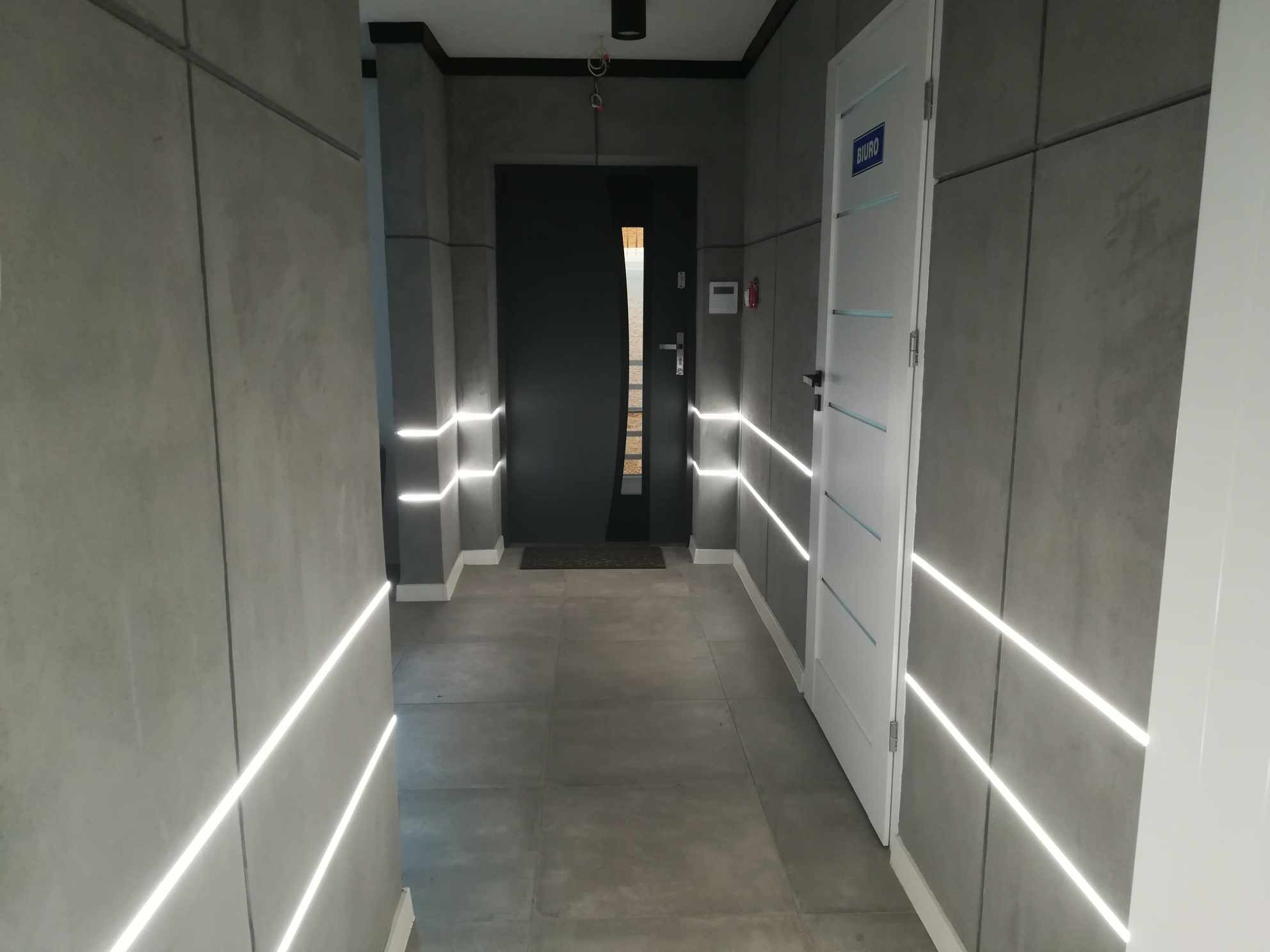 Nowoczesny korytarz biurowy z szarymi panelami ściennymi i liniowym oświetleniem LED. Widoczne drzwi wejściowe i drzwi do biura.
