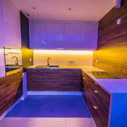 Nowoczesna kuchnia z białymi górnymi szafkami i drewnopodobnymi dolnymi, podświetlona ledami w kolorach fioletu i żółci, z blatem roboczym w kształcie litery L i drewnianą ścianą.