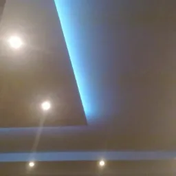 Nowoczesny sufit podwieszany z oświetleniem LED w kolorze niebieskim i punktowymi lampami halogenowymi, widok z dołu, fragment drewnianej framugi drzwi po lewej stronie.