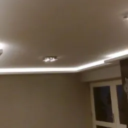 Nowoczesne oświetlenie sufitowe z listwami LED, podkreślające gładką powierzchnię sufitu podwieszanego w odcieniach beżu, z widocznymi dwoma lampami sufitowymi i fragmentem szafy oraz zegara...