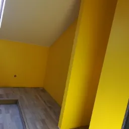 Żółte ściany i jasny sufit w pokoju na poddaszu z drewnianą podłogą, widoczny fragment okna.