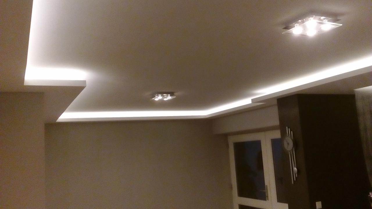 Nowoczesne oświetlenie sufitowe z listwami LED, podkreślające gładką powierzchnię sufitu podwieszanego w odcieniach beżu, z widocznymi dwoma lampami sufitowymi i fragmentem szafy oraz zegara...