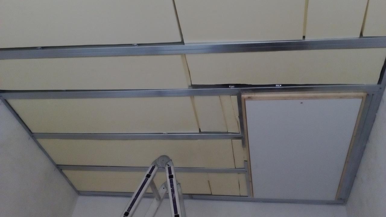 Montaż sufitu podwieszanego z widoczną konstrukcją metalową, płytami izolacyjnymi i klapą rewizyjną, z częściowo widoczną drabiną w rogu pomieszczenia.