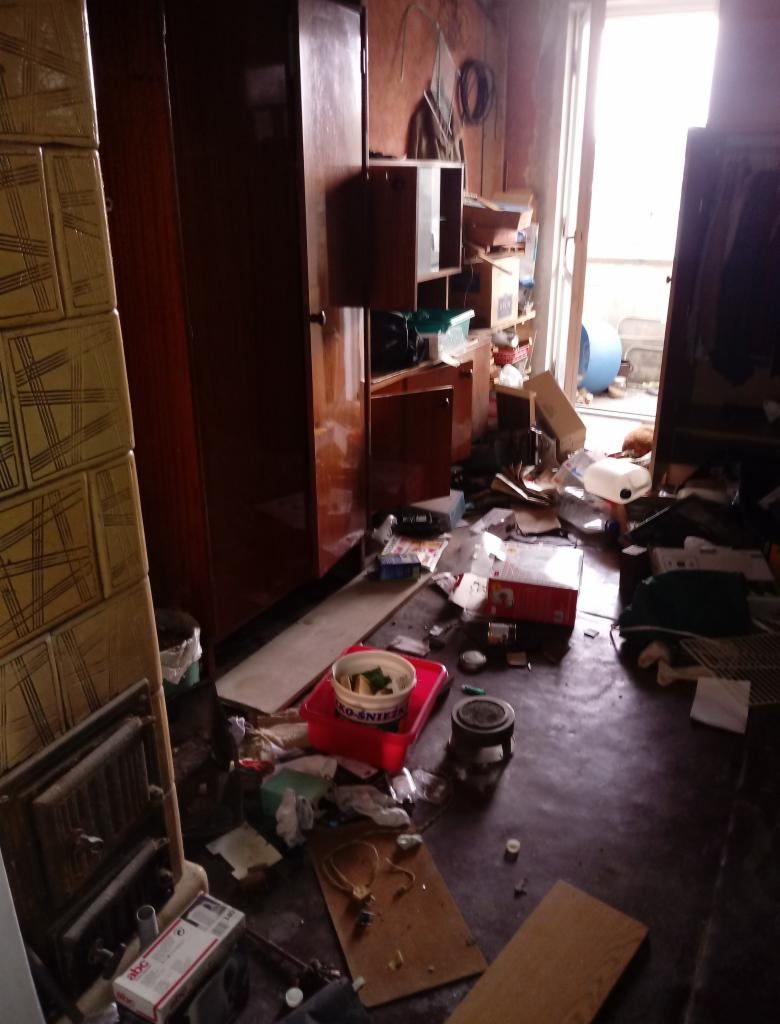 Zaniedbane wnętrze pokoju z widocznym bałaganem, stare meble, śmieci na podłodze i przybrudzona podłoga.