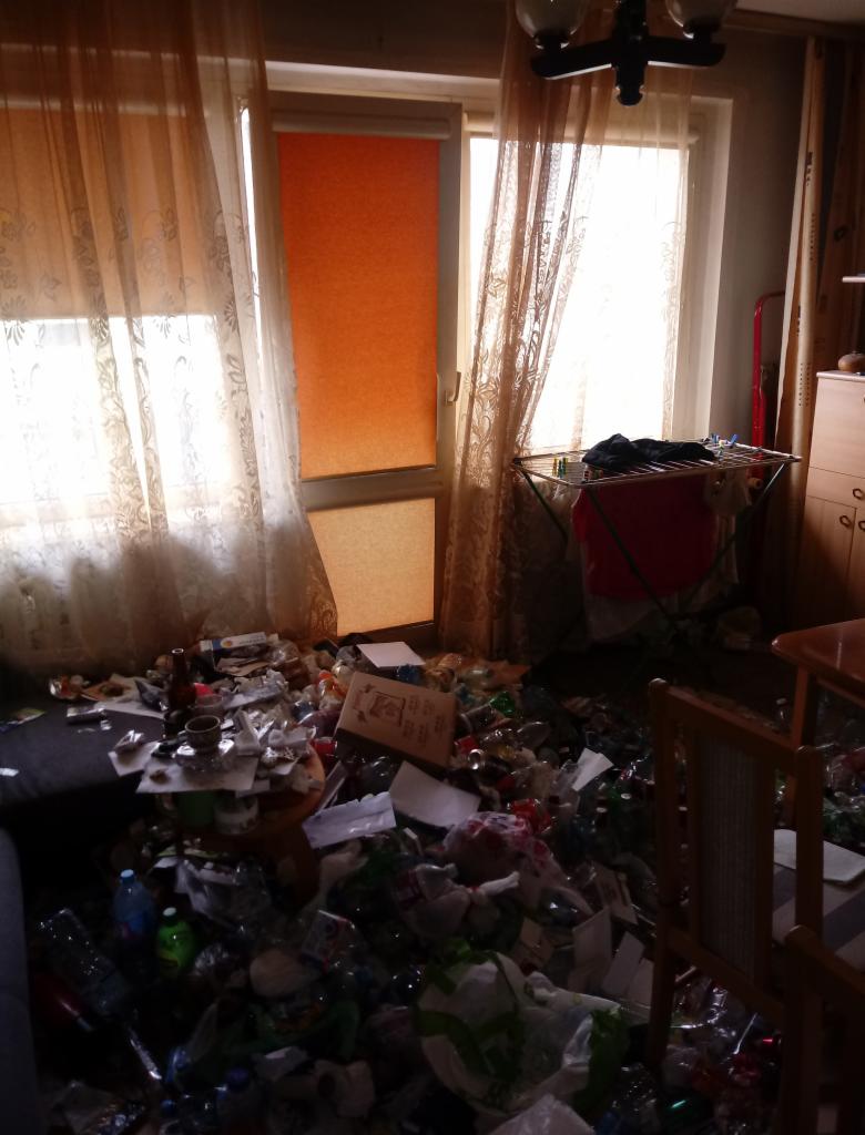 Zaniedbane wnętrze pokoju z nagromadzeniem śmieci i odpadów na podłodze, widoczne brudne zasłony i suszarka na pranie w tle.