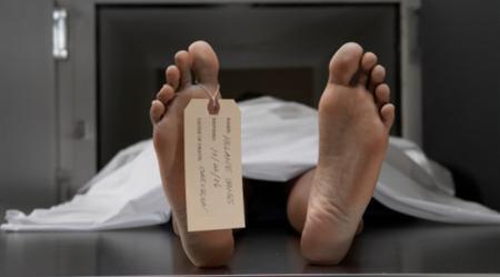 Stopy osoby z przywieszoną kartką identyfikacyjną, leżące na metalowym stole pod białym prześcieradłem, w tle ciemne pomieszczenie.