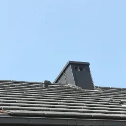 Nowoczesny dach z ciemnoszarej dachówki ceramicznej z kominem wentylacyjnym i elementami wentylacji połaciowej na tle błękitnego nieba.