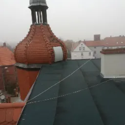 Widok z góry na pokrycie dachu z papy termozgrzewalnej, z kominem i wieżyczką z czerwonej cegły w tle zabudowa miejska w mglisty dzień.