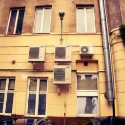 Fasada budynku z kilkoma zewnętrznymi jednostkami klimatyzacji zamontowanymi nad oknami, widoczny również pionowy metalowy przewód wentylacyjny.