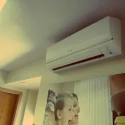 Biały klimatyzator ścienny zamontowany w pomieszczeniu, widoczny nad lustrem z odbiciem osób.