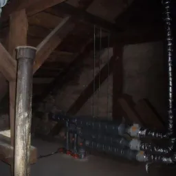 Instalacja rur w ciemnym poddaszu z drewnianą konstrukcją, widoczne rury izolowane termicznie i fragment instalacji z zaworami.