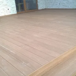 Nowo ułożony taras z desek kompozytowych w odcieniach brązu, z widocznymi podporami i fragmentem elewacji z białej cegły.