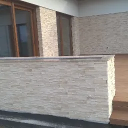 Wykończona elewacja domu jednorodzinnego z dekoracyjnym kamieniem elewacyjnym w odcieniach beżu, widoczny fragment tarasu z desek kompozytowych i okna z drewnianą stolarką.