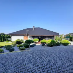 Nowoczesny dom jednorodzinny w Głuszynie z ciemnym dachem, szarą elewacją i ozdobnym ogrodem z kamienną ścieżką i licznymi krzewami.