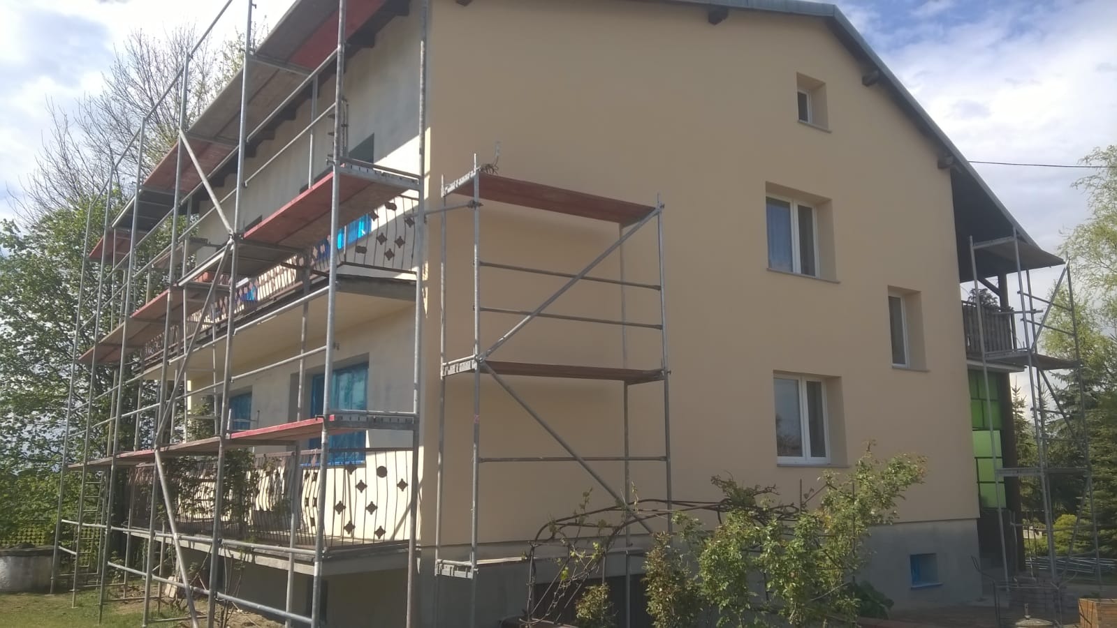 Dwukondygnacyjny dom jednorodzinny w trakcie prac elewacyjnych, otoczony rusztowaniem; świeżo otynkowana, beżowa elewacja i częściowo zasłonięte okna folią ochronną.