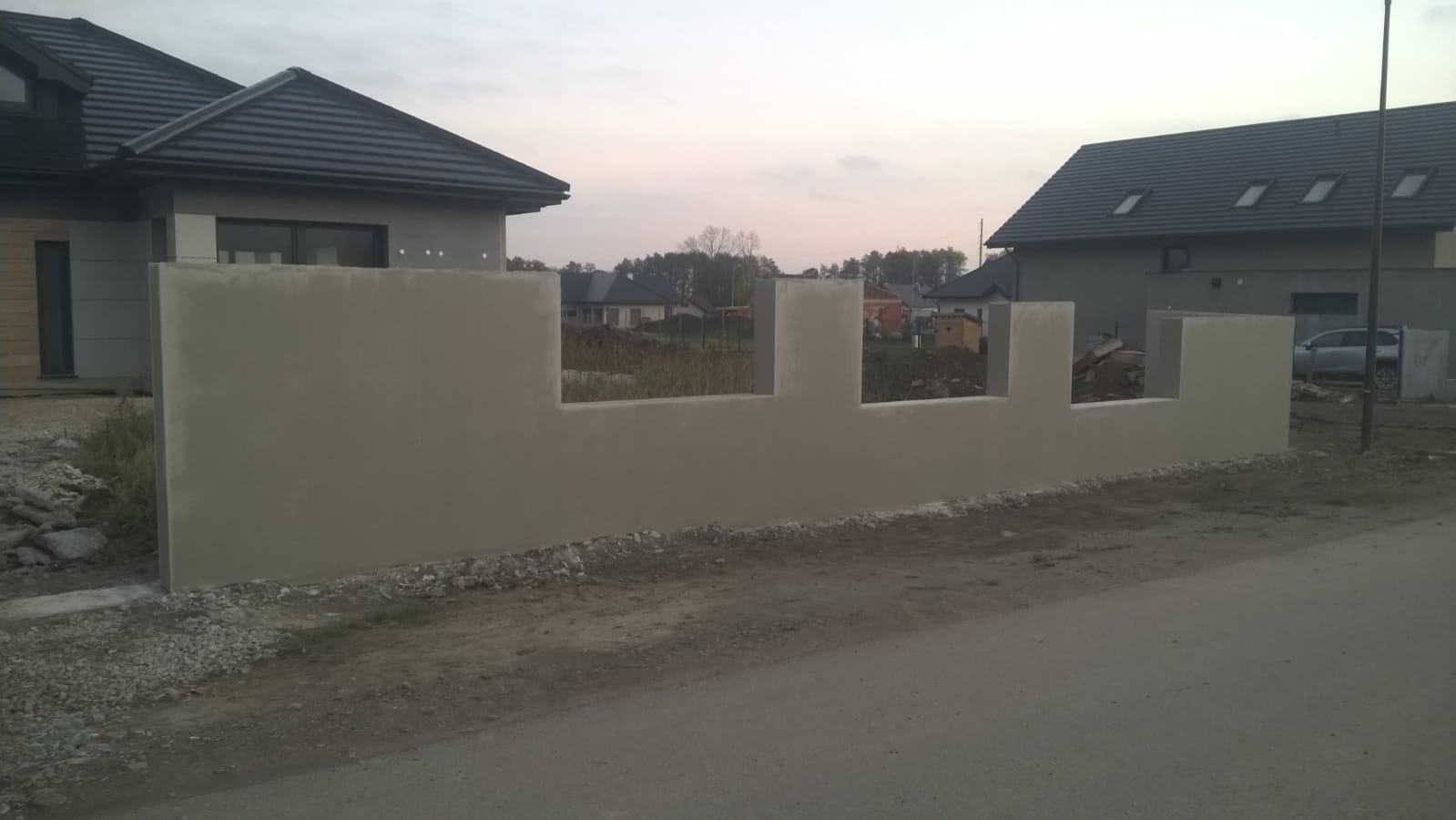 Nowo wybudowany, surowy mur z trzema prostokątnymi prześwitami, widoczny na tle domów jednorodzinnych w spokojnej okolicy.