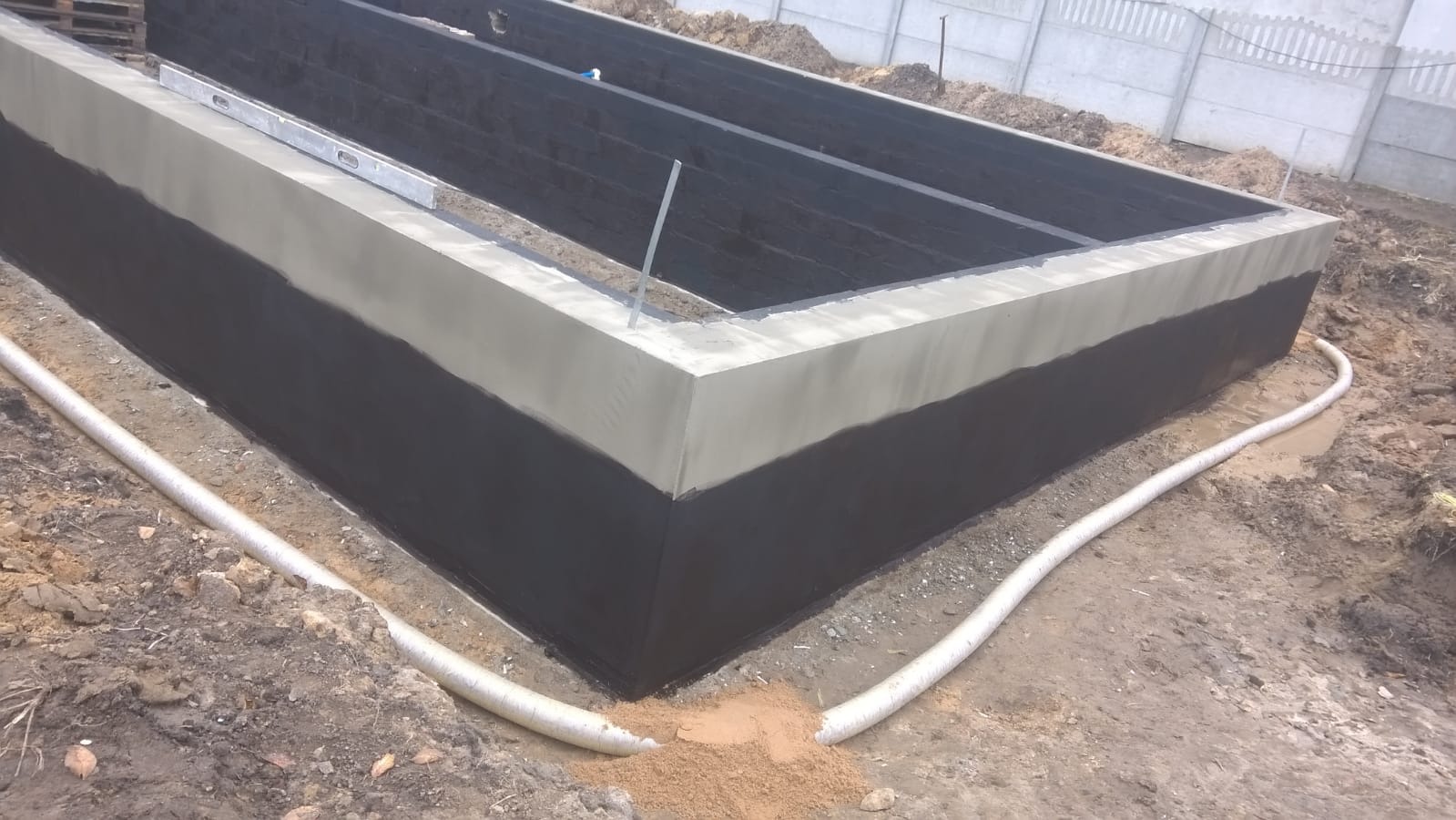 Wykop pod fundamenty domu z hydroizolacją i wylewką z betonu, widoczny poziomica i biała rura drenażowa.