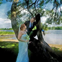 Para młoda w Suwałkach pozuje przy starym, rozłożystym drzewie nad brzegiem jeziora, panna młoda w białej sukni trzyma bukiet, pan młody w garniturze siedzi na konarze.
