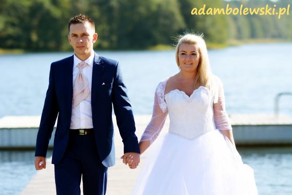 Para młoda, on w granatowym garniturze z beżowym krawatem, ona w białej sukni ślubnej z koronkowymi rękawami, spacerują trzymając się za ręce po drewnianym pomoście nad jeziorem w słoneczny dzień.