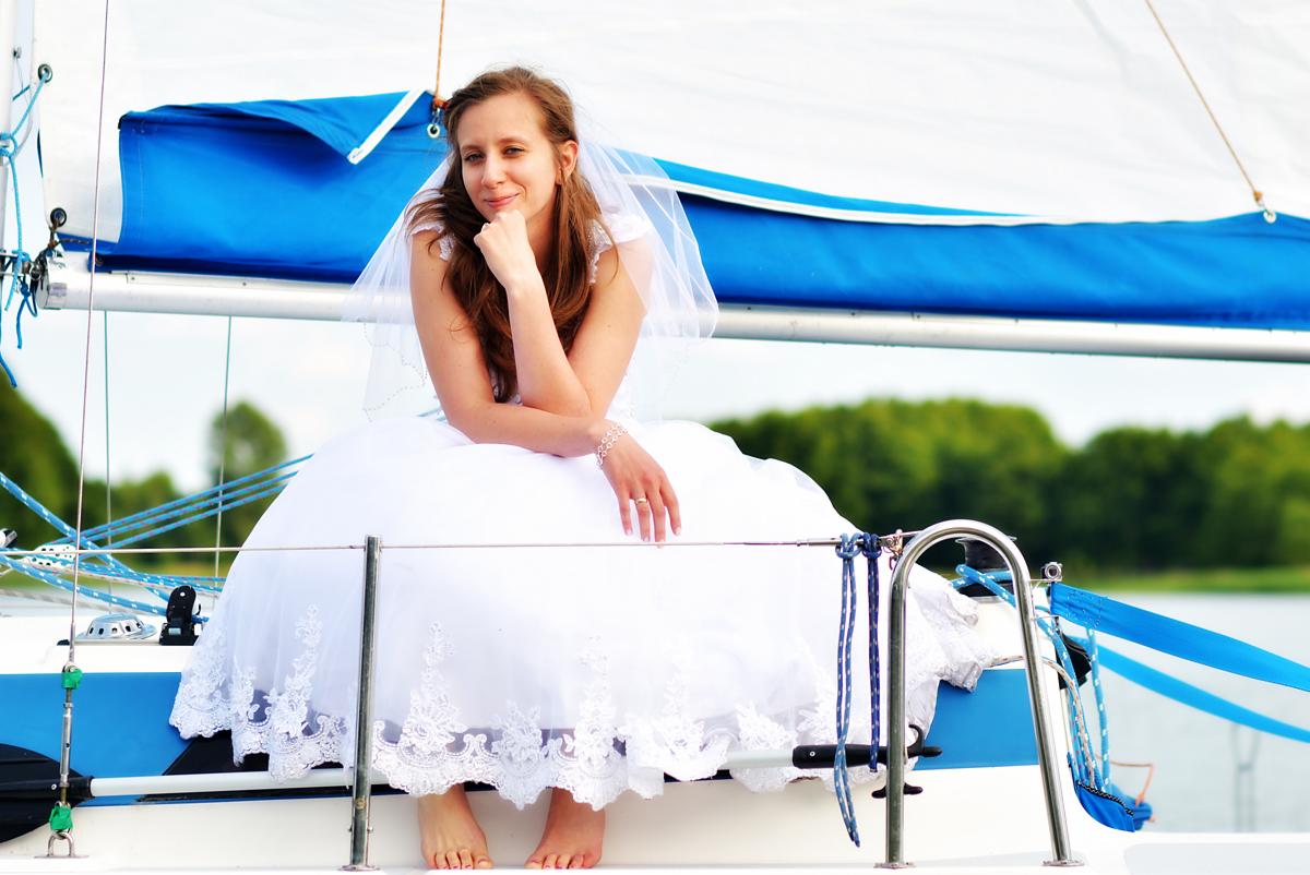 Panna młoda w białej sukni i welonie siedzi na jachcie z niebieskimi elementami, uśmiecha się, w tle woda i zieleń.