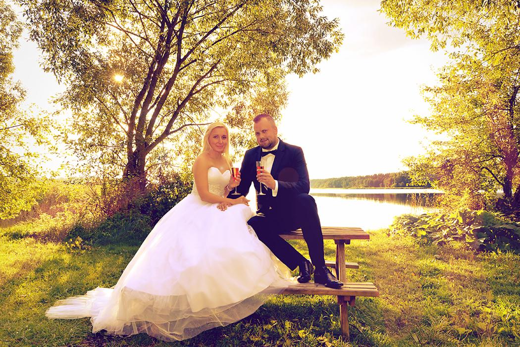 Para młoda siedząca na drewnianej ławce w plenerze, toast szampanem na tle jeziora i zachodzącego słońca przebijającego przez korony drzew.