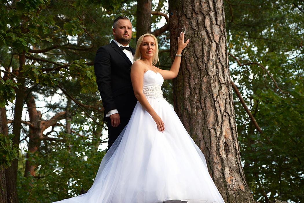 Para młoda w plenerze: panna młoda w białej sukni bez ramiączek opiera się o sosnę, obok pan młody w czarnym smokingu z muchą, w tle zielone drzewa.