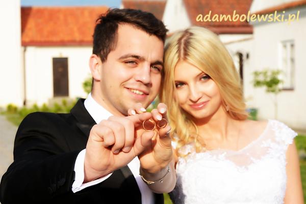 Uśmiechnięta para młoda prezentuje złote obrączki na tle białego budynku z pomarańczowym dachem.