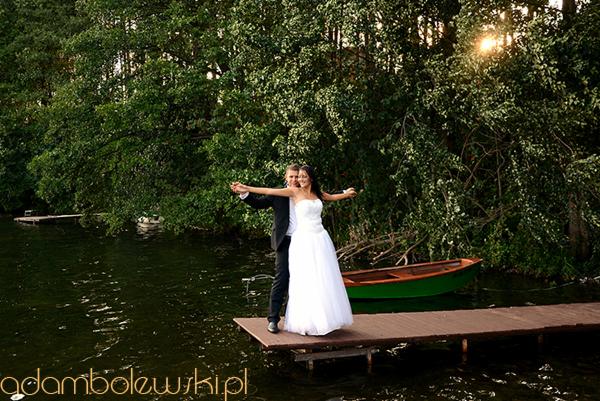 Para młoda na drewnianym pomoście nad jeziorem, pozująca z rozłożonymi ramionami, w tle zielony las i łódka.