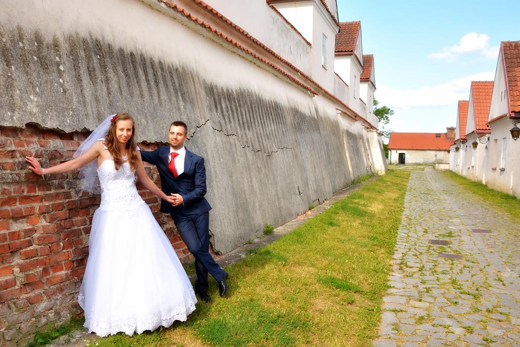 Para młoda pozuje przy ceglanej ścianie z pęknięciami, on w granatowym garniturze z czerwoną koszulą, ona w białej sukni ślubnej z welonem, w tle brukowana droga i budynki z czerwonymi dachami.