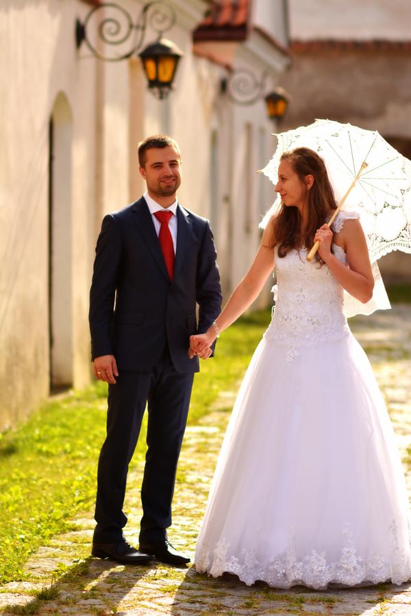 Para młoda trzymająca się za ręce na brukowanej uliczce, on w granatowym garniturze i czerwonym krawacie, ona w białej sukni z parasolką, w tle budynki z lampami.