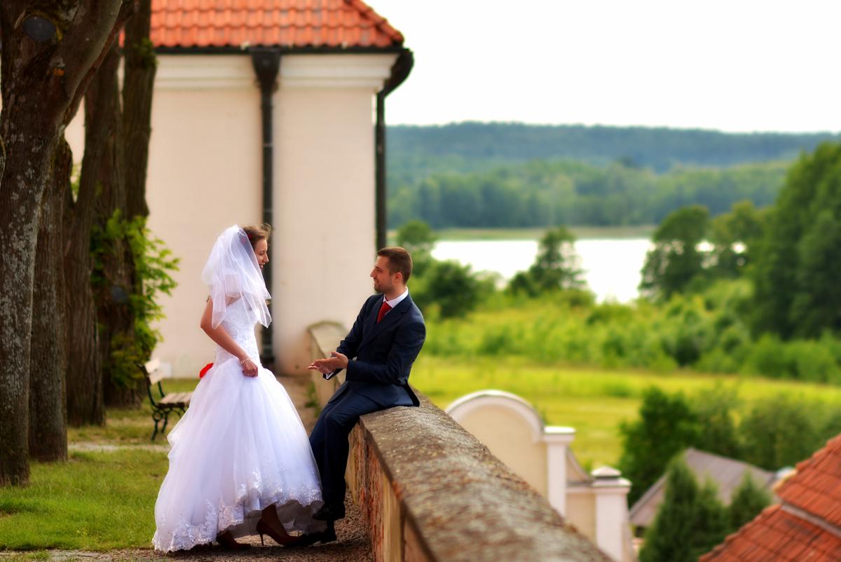 Para młoda w plenerze: panna młoda w białej sukni z welonem i czerwonym akcentem, pan młody w granatowym garniturze z czerwoną koszulą, siedzący na murku z widokiem na jezioro i zieleń.