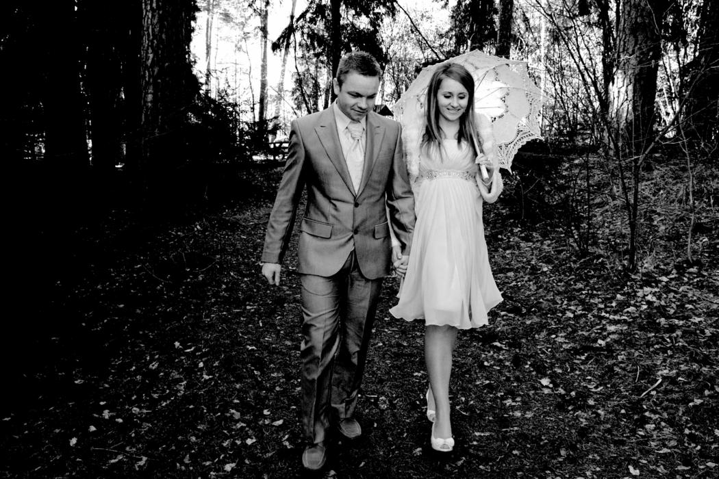 Para młoda w lesie, czarno-białe zdjęcie: mężczyzna w garniturze i kobieta w jasnej sukience trzymająca koronkowy parasol, spacerują po leśnej ścieżce.