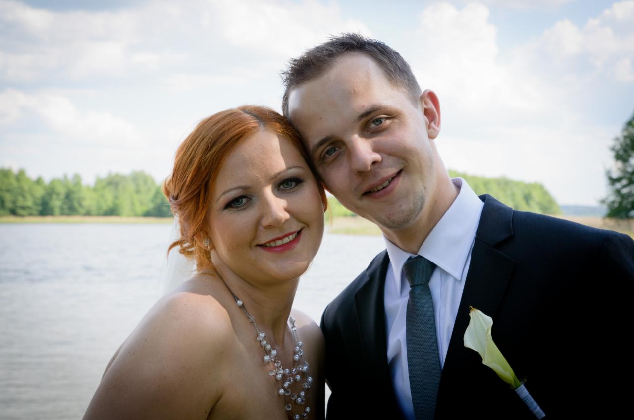 Uśmiechnięta para młoda, on w czarnym garniturze z białą kalą w butonierce, ona z rudymi włosami i perłowym naszyjnikiem, pozuje na tle jeziora i zieleni.