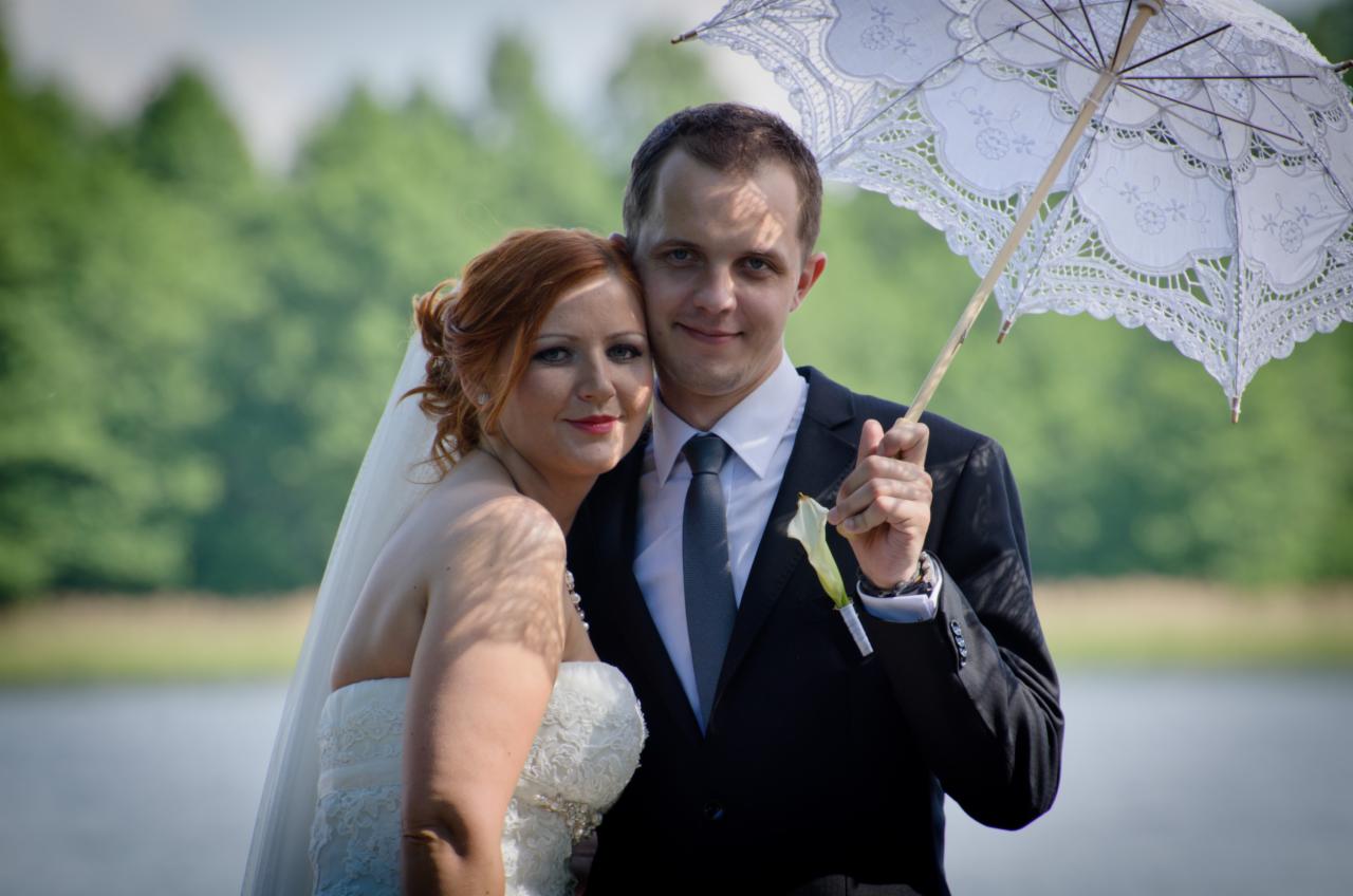 Uśmiechnięta para młoda, on w czarnym garniturze z butonierką i zegarkiem, ona w białej sukni z koronką i welonem, pozuje z białym, koronkowym parasolem na tle zieleni i wody.