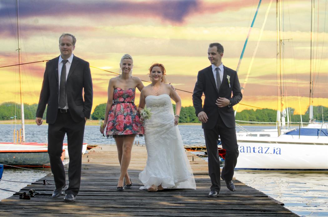 Para młoda i goście weselni spacerują po drewnianym molo nad jeziorem o zachodzie słońca, z żaglówkami w tle.