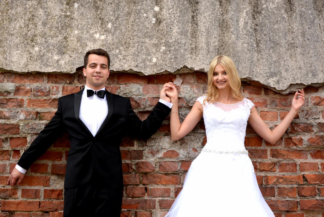 Uśmiechnięta para młoda, on w czarnym smokingu i muszce, ona w białej sukni ślubnej z koronkową górą i zdobionym paskiem, trzymają się za ręce na tle ceglanej ściany z szarym, popękanym tynkiem u...