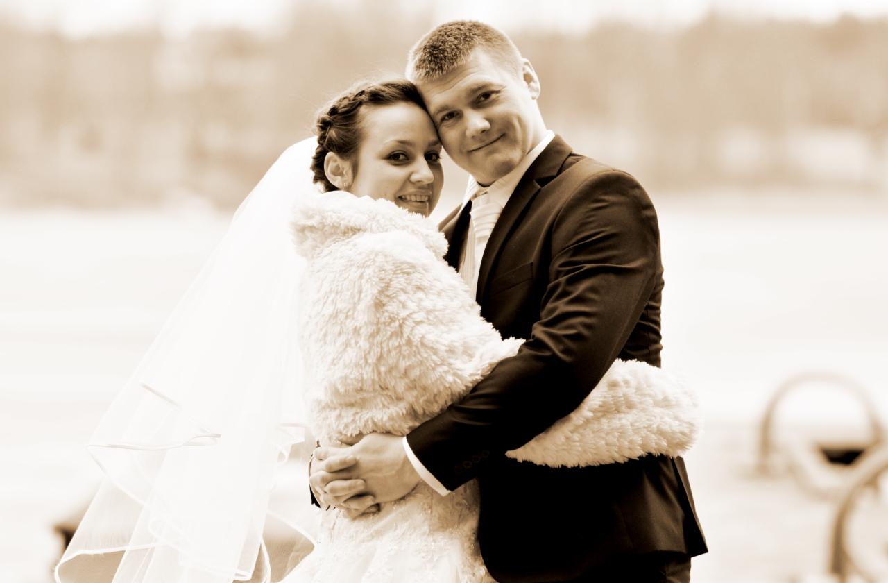 Uśmiechnięta para młoda w objęciach, panna młoda w futrzanym bolerku i welonie, pan młody w ciemnym garniturze, sepia.