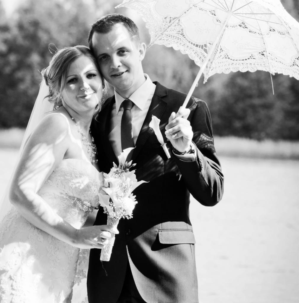 Czarno-białe zdjęcie uśmiechniętej pary młodej: panna młoda w koronkowej sukni z bukietem kalii, pan młody w garniturze, trzyma koronkowy parasol nad głową, w tle woda i drzewa.