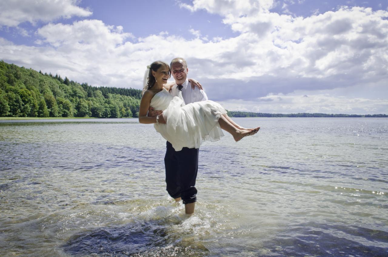 Uśmiechnięty pan młody niesie pannę młodą w sukni ślubnej przez płytką wodę jeziora, w tle las i pochmurne niebo.