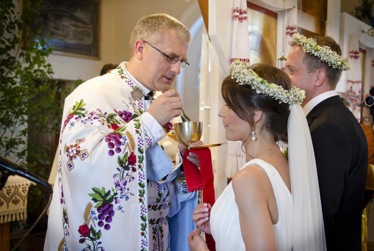 Ksiądz w bogato haftowanej szacie podaje komunię pannie młodej w wianku z białych kwiatów podczas ceremonii ślubnej.