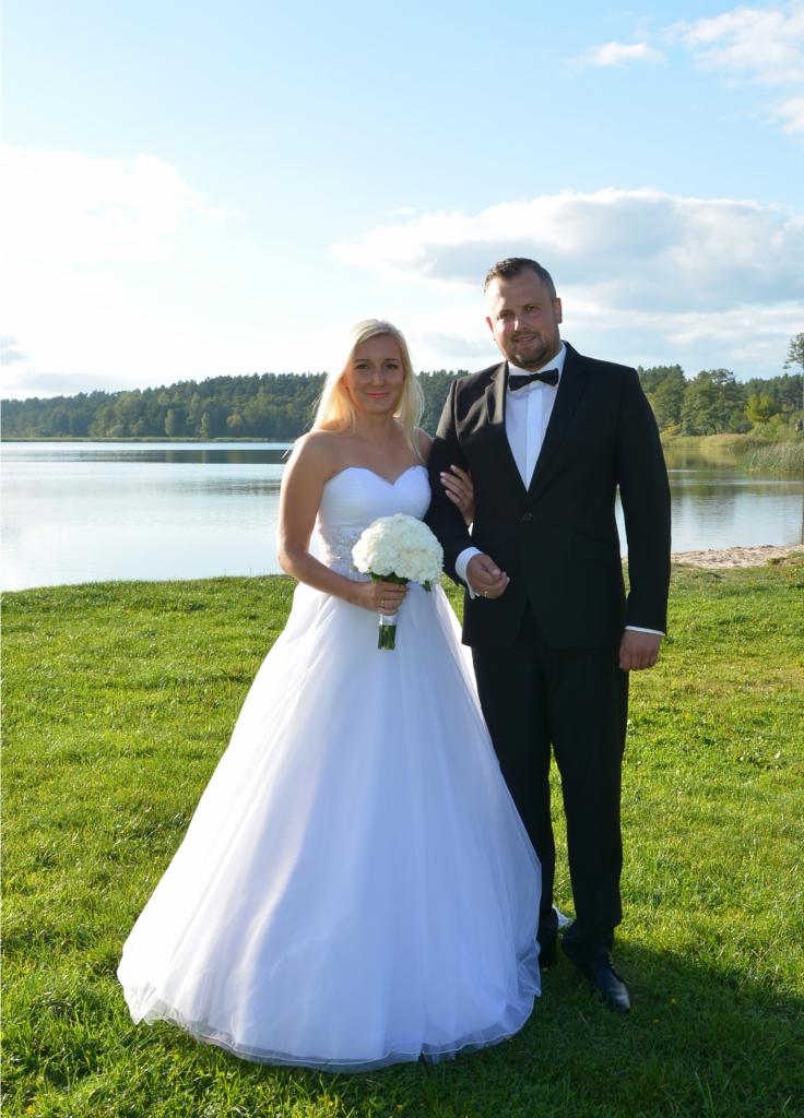 Para młoda w plenerze nad jeziorem: panna młoda w białej sukni z bukietem białych róż, pan młody w czarnym garniturze z muchą, oboje stoją na zielonej trawie.