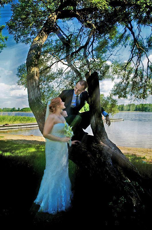 Para młoda w Suwałkach pozuje przy starym, rozłożystym drzewie nad brzegiem jeziora, panna młoda w białej sukni trzyma bukiet, pan młody w garniturze siedzi na konarze.