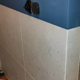 Fragment łazienki z wykończoną ścianą: dolna część z jasnymi, kwadratowymi płytkami imitującymi kamień, górna część pomalowana na niebiesko. Na ścianie widoczny czarny wieszak i osprzęt.