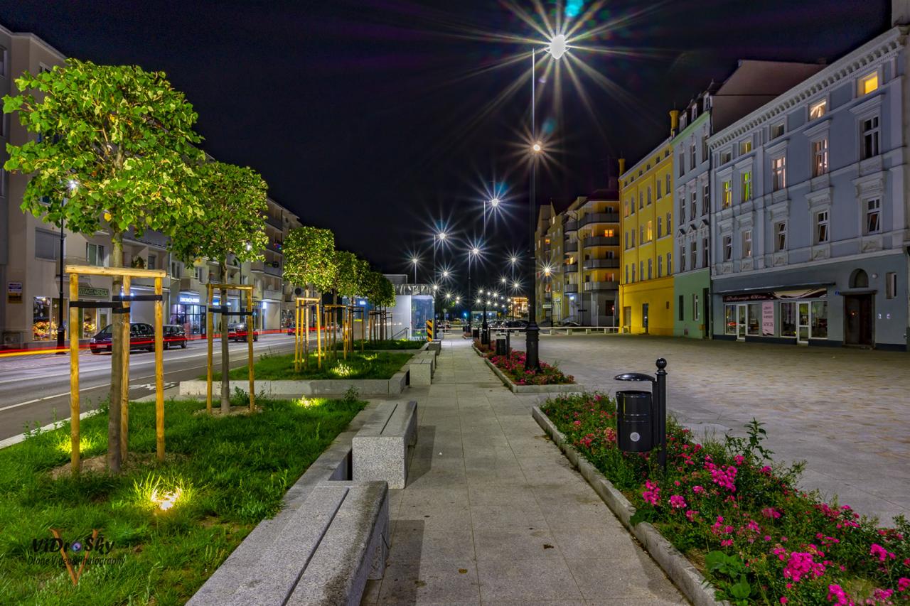 Nocny widok na oświetloną ulicę miejską z rzędem drzew, granitowymi ławkami, kwietnikami z różowymi kwiatami i kolorowymi budynkami w tle.