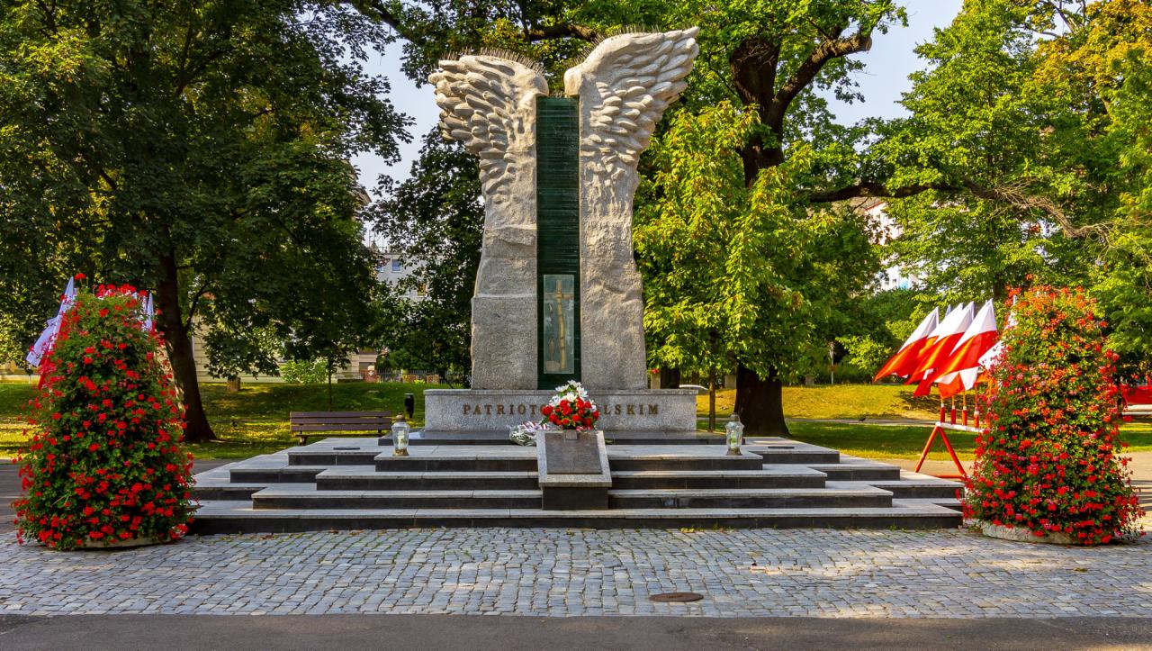 Pomnik w formie rozpostartych skrzydeł z napisem 'Patriotom Polskim', otoczony kwiatami i polskimi flagami w parku.
