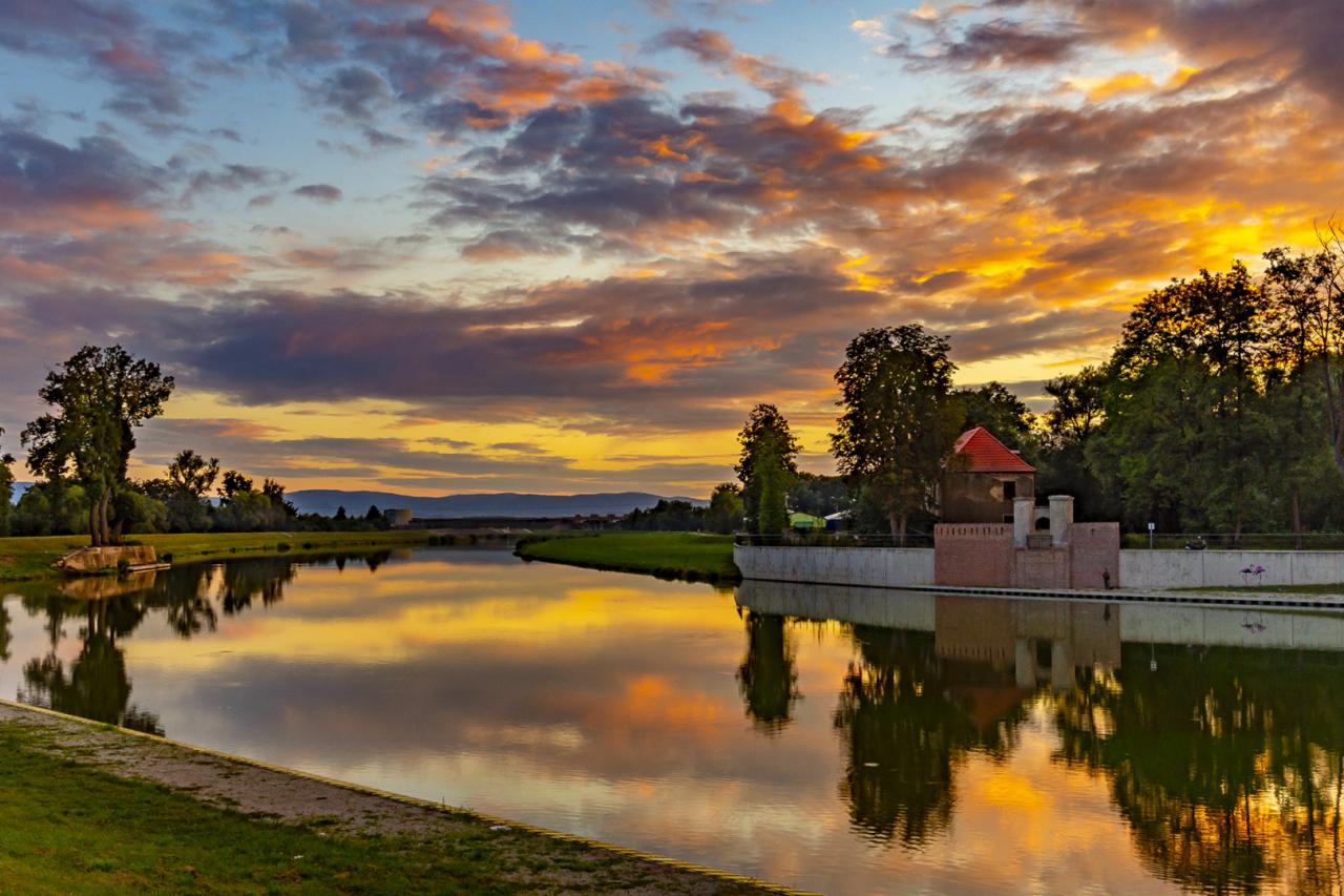 Spokojna rzeka odbija dramatyczne, pomarańczowo-szare chmury o zachodzie słońca; w tle widoczny mur z cegły z wieżyczką i drzewa