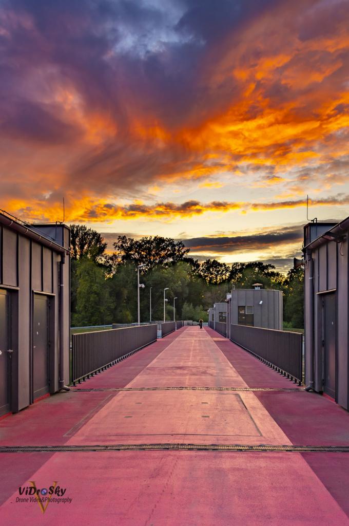 Czerwony chodnik na moście z szarymi budynkami po bokach, widok na zachodzące słońce i drzewa w tle.