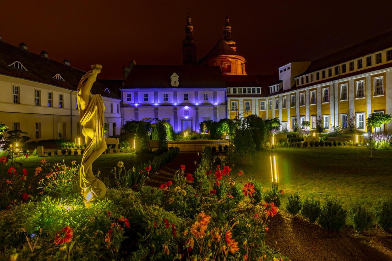 Nocny widok na barokowy pałac z ogrodem, oświetlony ciepłym światłem, z rzeźbą w pierwszym planie i kwiatami w odcieniach czerwieni i żółci.