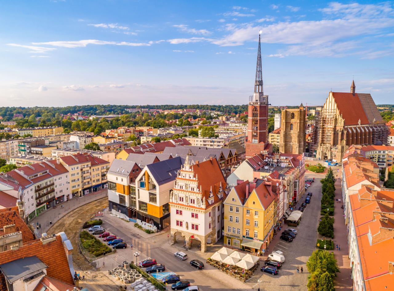 Panorama miasta z lotu ptaka, widoczne zabytkowe budynki, kościoły z wieżami, czerwone dachy i nowoczesna architektura w słoneczny dzień.