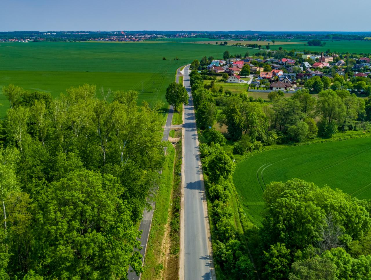 Panoramiczny widok z lotu ptaka na wiejską drogę, otoczoną polami i lasami, prowadzącą do małej miejscowości z czerwonymi dachami.